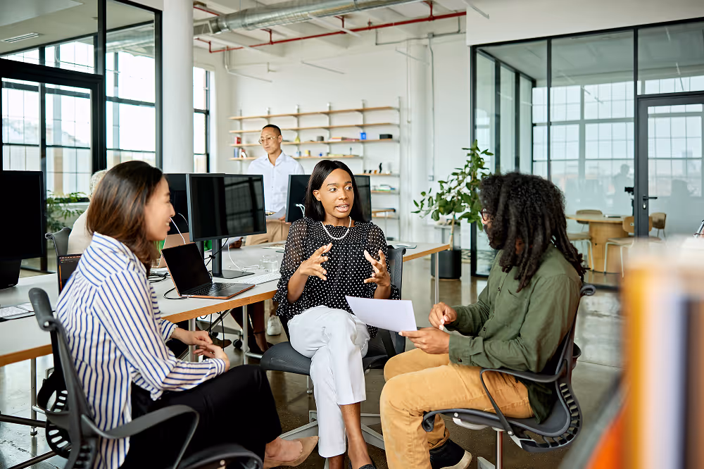 Three people seated and having a discussion in a modern office with computers and shelves in the background.