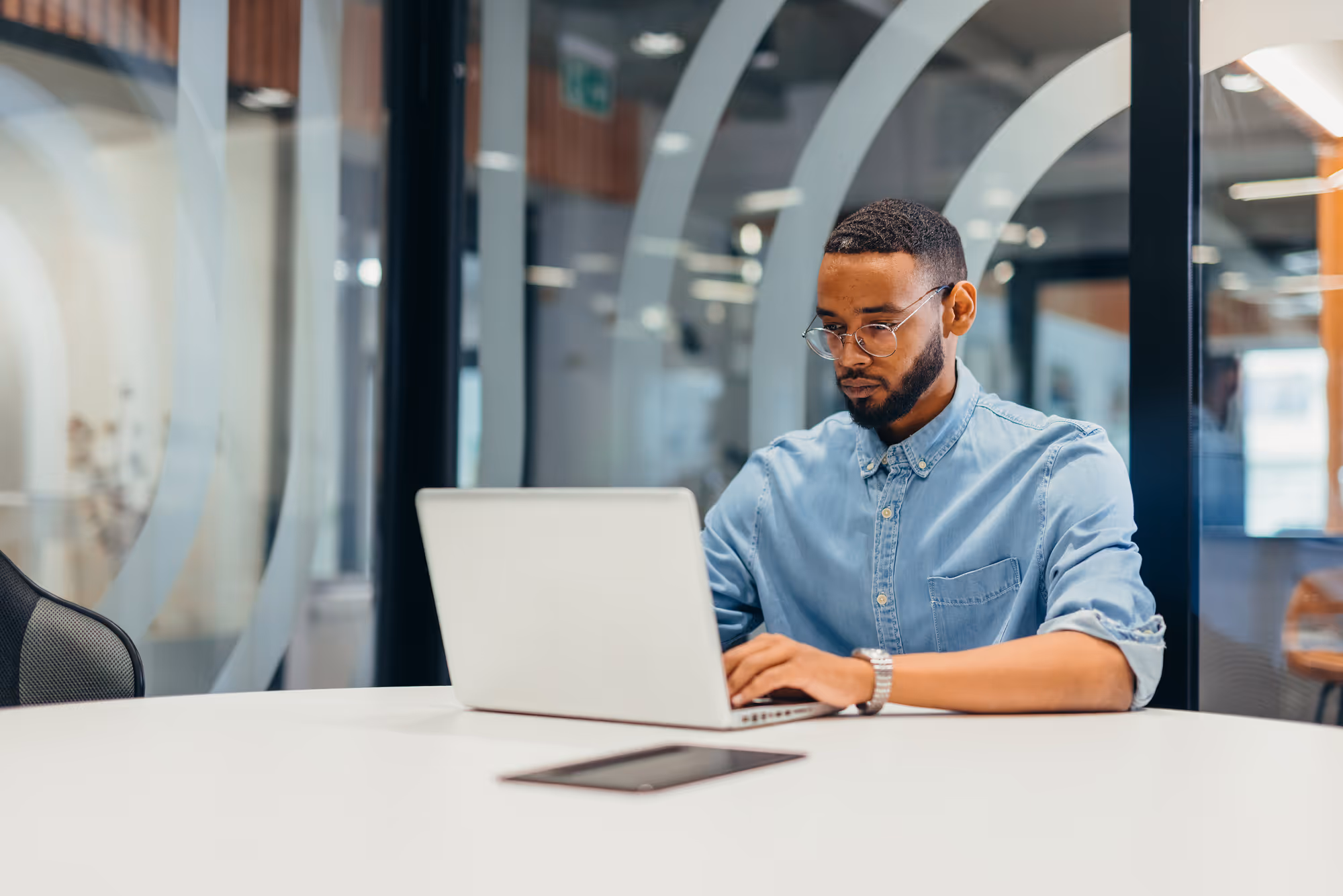 Man with glasses and beard working on a laptop at a white table in a modern office.