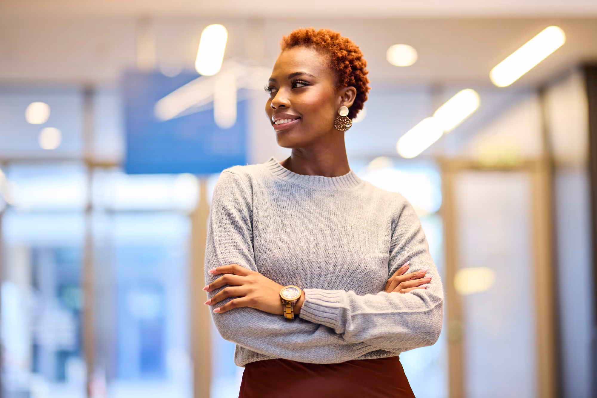 Confident woman with short curly hair wearing a grey sweater and watch, standing with arms crossed in a well-lit office.