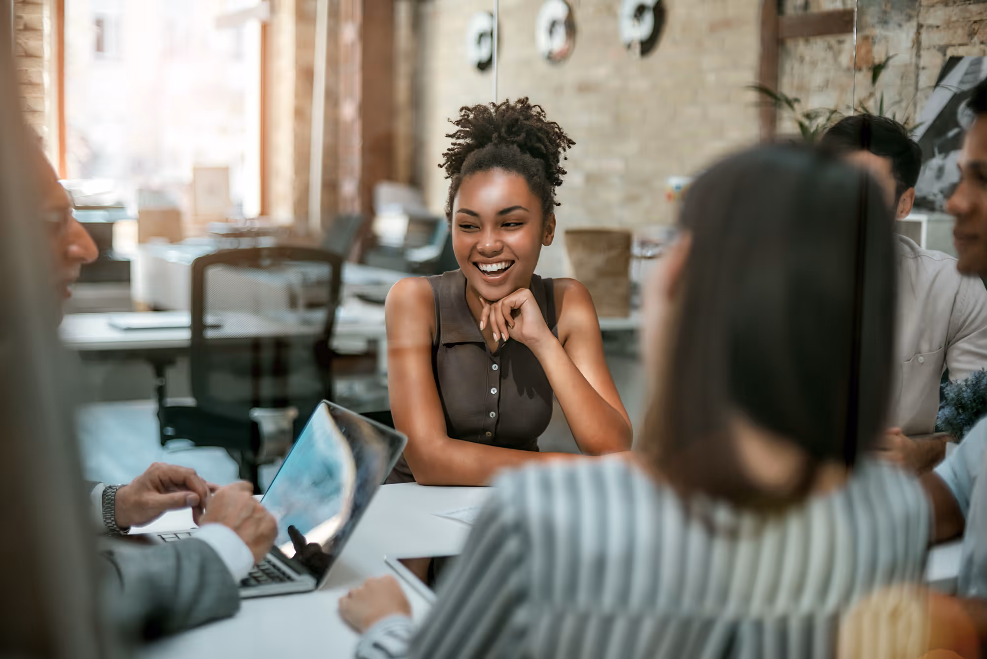 Smiling young woman with curly hair sitting at a table with colleagues in a modern office.