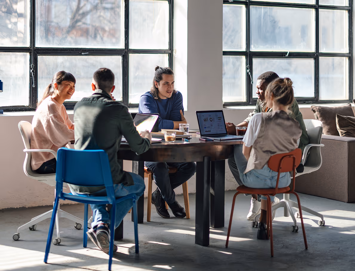 Five diverse young adults sitting around a table with laptops, engaged in a meeting in a bright room with large windows.