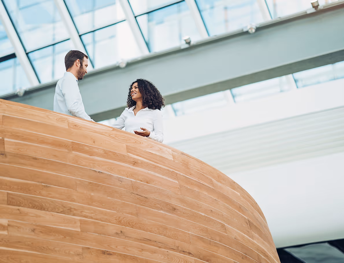 A man and a woman having a friendly conversation while standing on a wooden balcony inside a modern building with large windows.