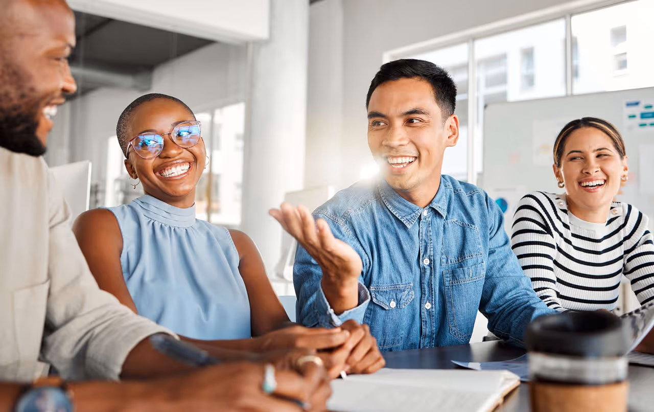 Four diverse colleagues smiling and engaging in a lively conversation at a modern office table.