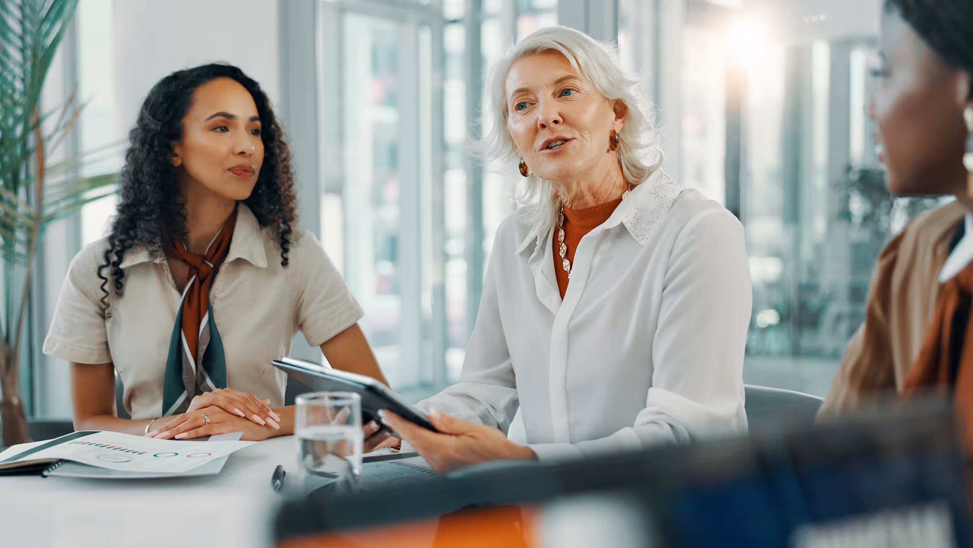 Three professional women in an office meeting, one holding a tablet and discussing while the others listen attentively.