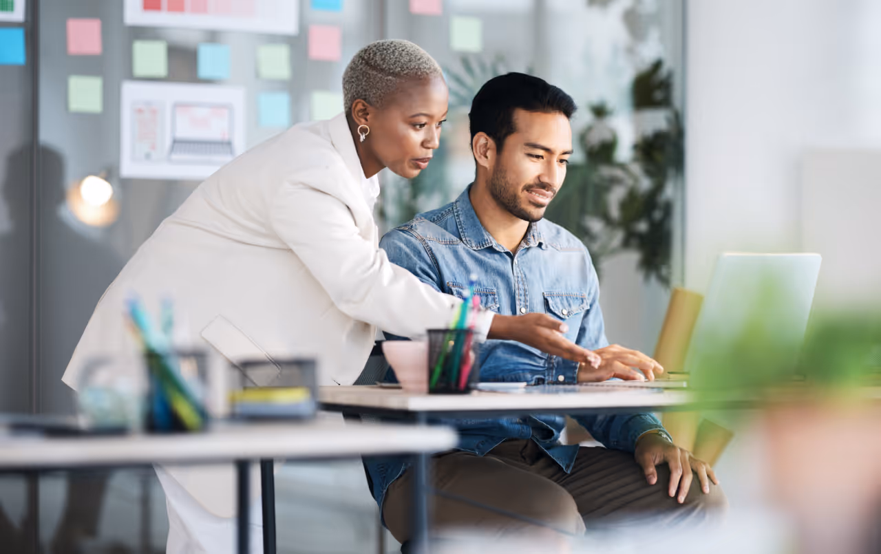 Woman in white blazer leaning over to explain something on a laptop to a seated man in a denim shirt in a brightly lit office.