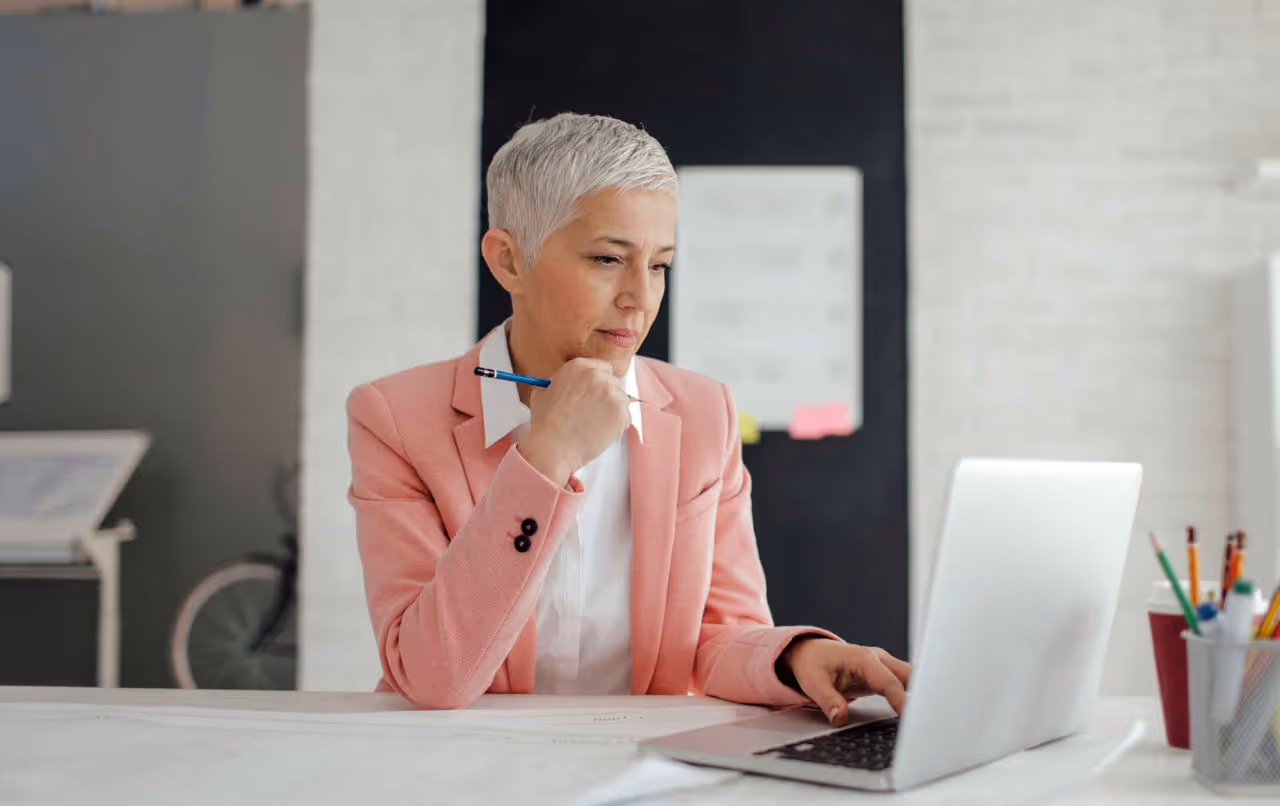 Woman with short gray hair in a pink blazer working thoughtfully on a laptop at a white desk.