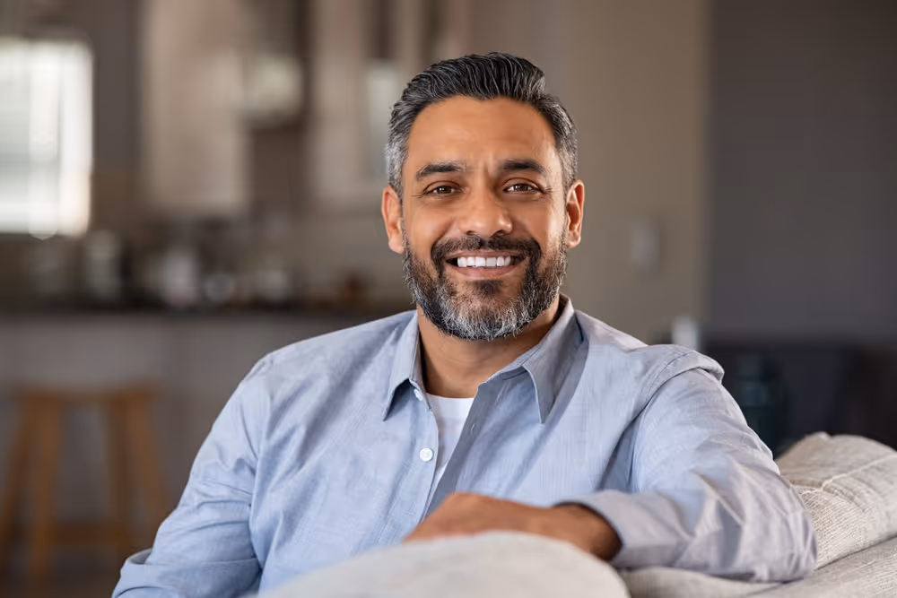 Smiling middle-aged man with salt-and-pepper beard sitting on a couch in a modern living room.