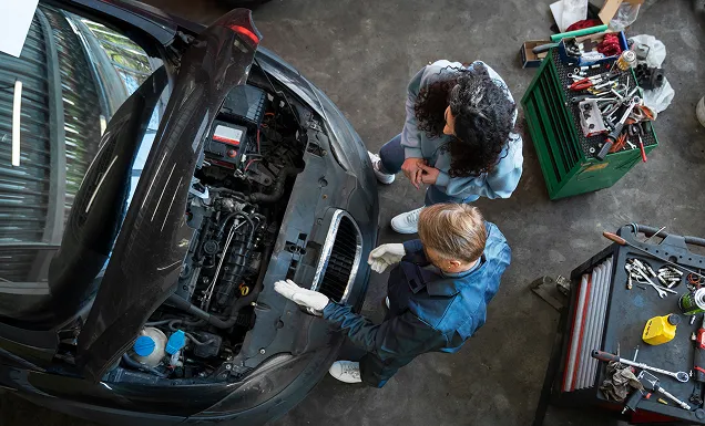 Mechaniker erklärt einer Frau den Motor eines schwarzen Autos in einer Werkstatt.