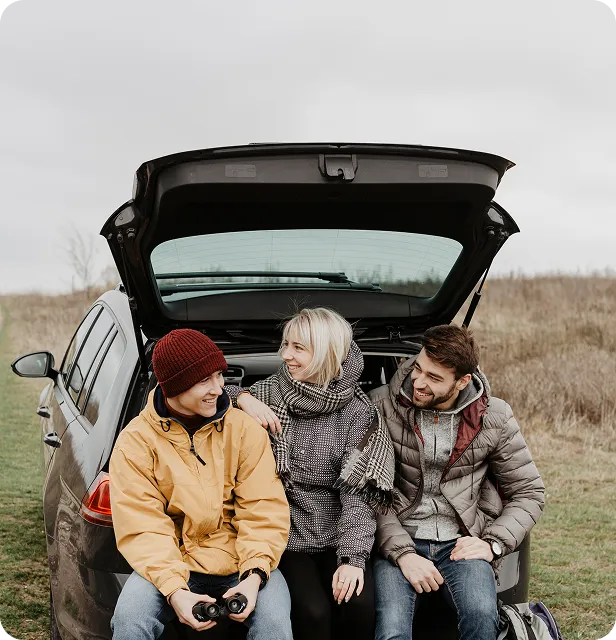 Drei junge Freunde sitzen im geöffneten Kofferraum eines Autos und unterhalten sich draußen auf einer Wiese.
