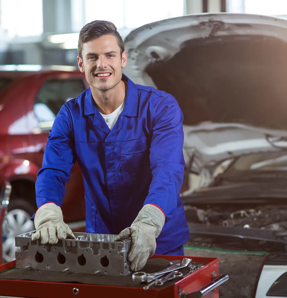 Ein Mechaniker in blauer Arbeitskleidung und Handschuhen hält ein Motorenteil in einer Werkstatt mit offenem Autohub.