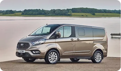 Brauner Kleinbus steht am Ufer vor ruhigem Wasser und bewaldeter Landschaft.