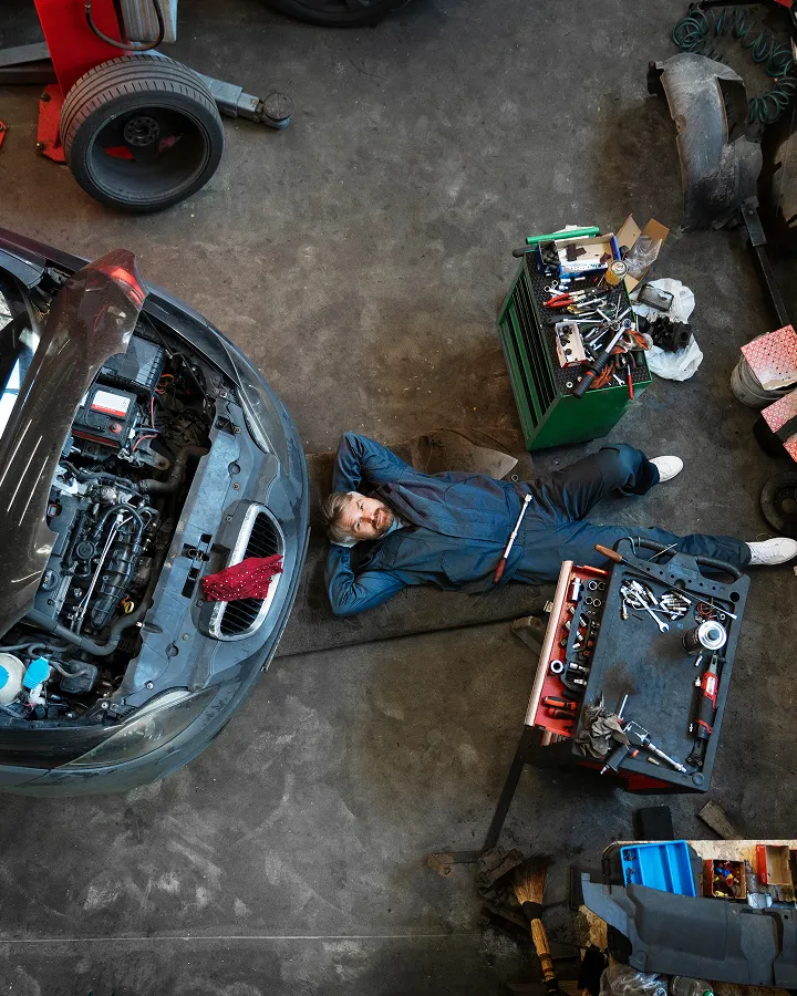 Mechaniker liegt entspannt auf einer Matte unter der Motorhaube eines Autos in einer Werkstatt mit Werkzeugen und Autoteilen um ihn herum.