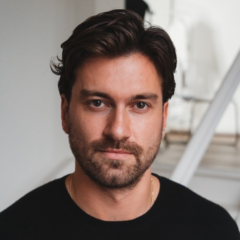Portrait of a man with dark hair and a beard wearing a black shirt, looking directly at the camera indoors.