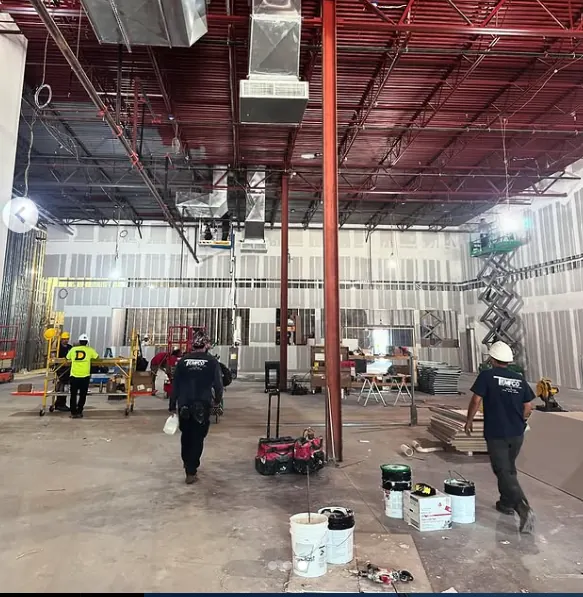 Interior of a large commercial building under construction with workers wearing hard hats and construction equipment scattered around.