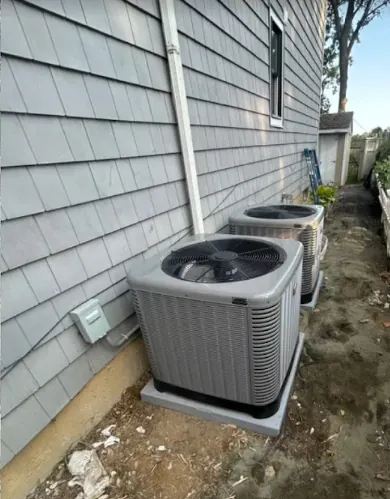 Two gray outdoor air conditioning units installed side by side next to a house with gray siding.