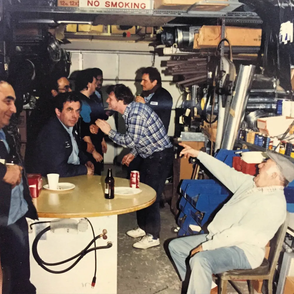 Group of men socializing in a workshop or garage with tools and equipment on shelves, one man seated pointing while others stand or sit around a table with drinks and plates.