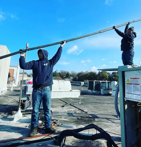 Two workers on a rooftop handling a long metal pipe, with one standing on the roof and the other on a rooftop unit under a bright blue sky.