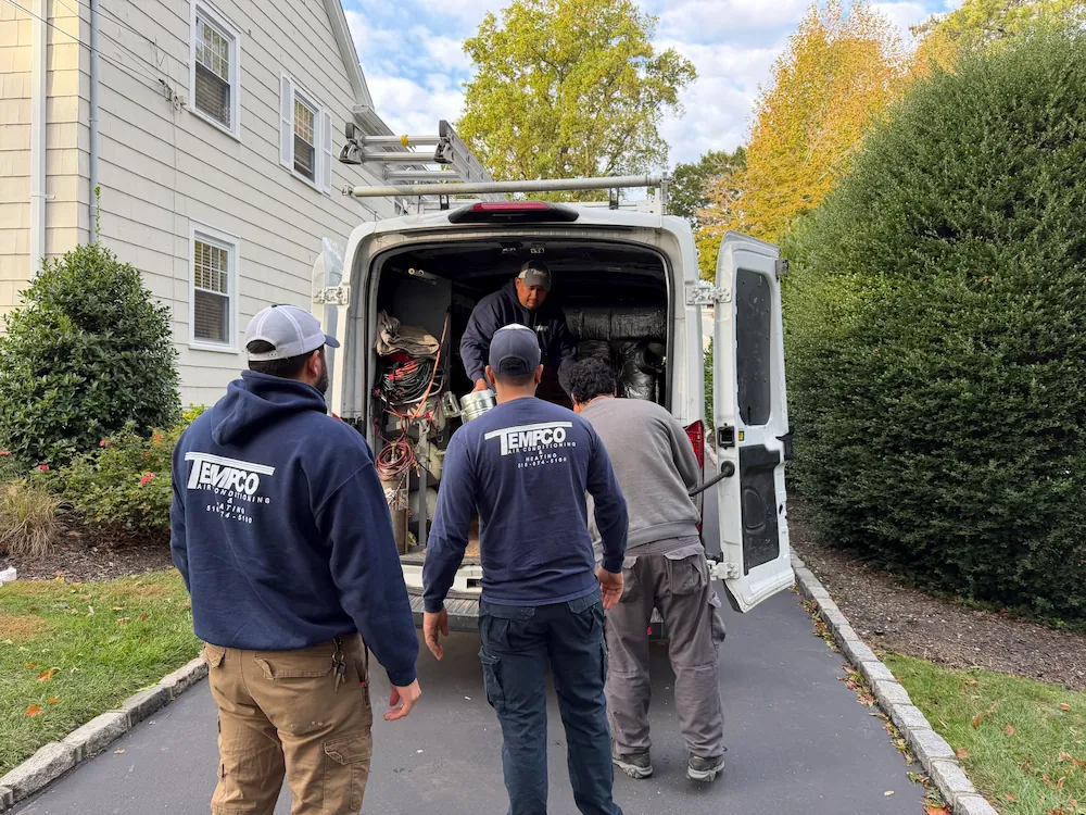 Three workers carrying equipment into the back of a white service van parked beside a house.