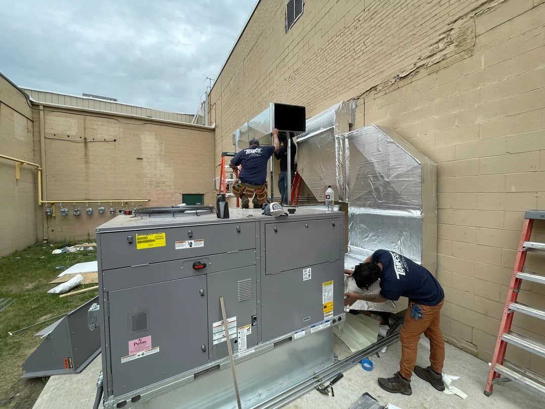 Three workers installing large HVAC ductwork and equipment outside a beige brick building.