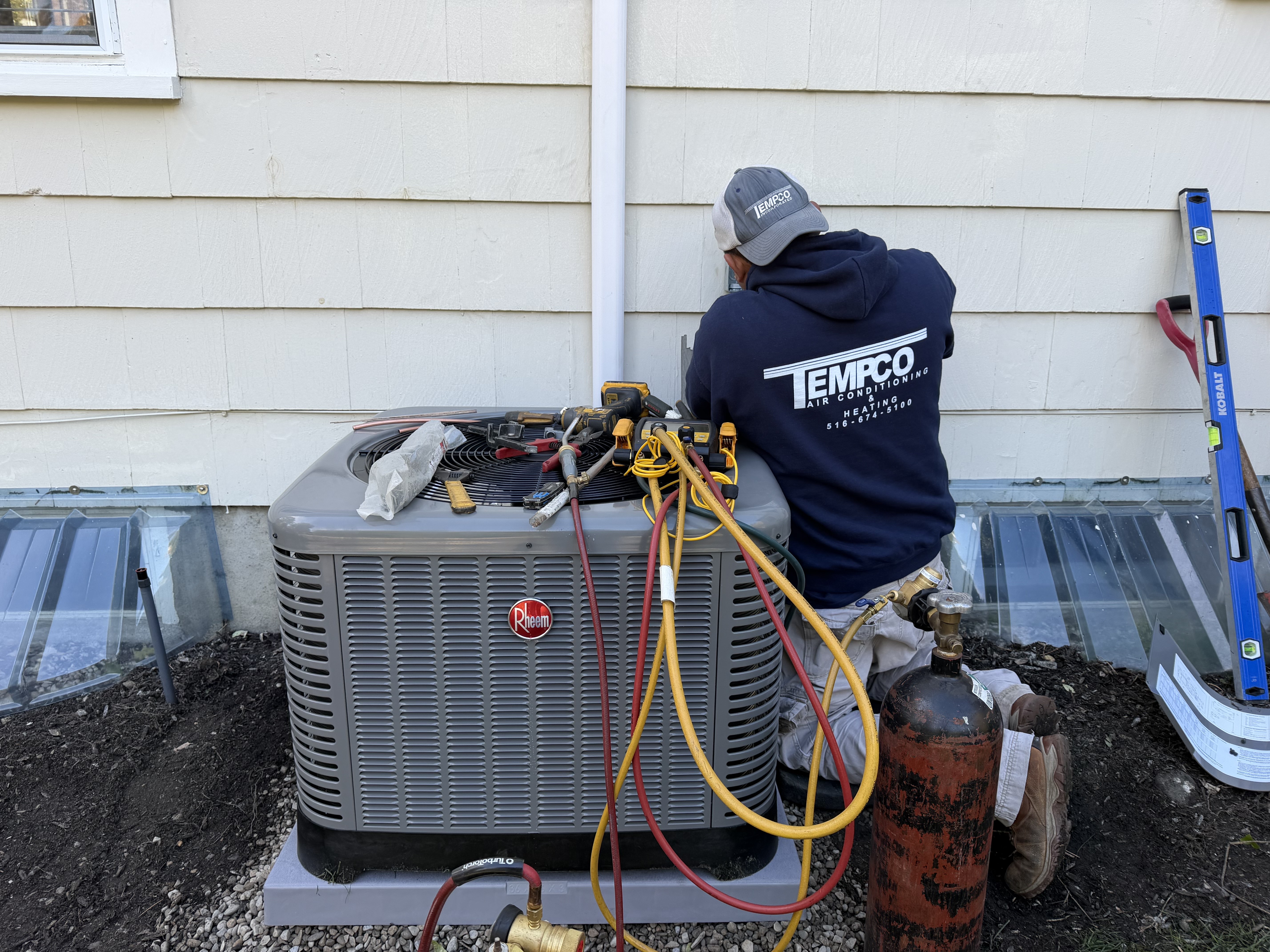 Technician wearing TEMPCO Air Conditioning hoodie working on a Rheem air conditioning unit outside a house.