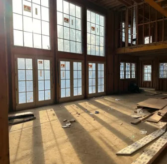 Sunlight streaming through large glass doors and windows in an unfinished wooden room with scattered construction materials on the floor.