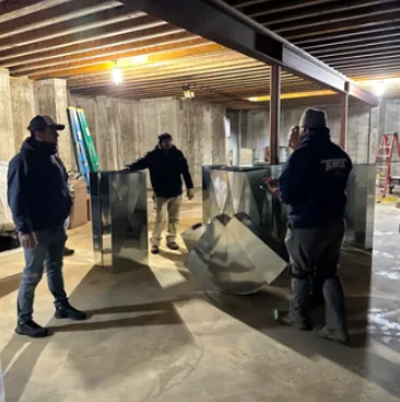 Four men standing around shiny metal ductwork pieces in an unfinished basement with exposed wooden beams and concrete walls.