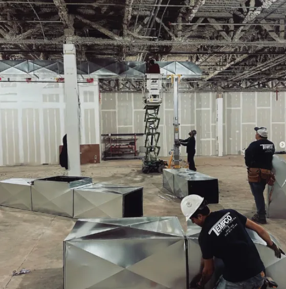 Construction workers installing large metal HVAC ductwork inside a building with exposed ceiling.