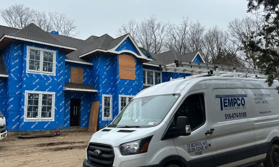 Two-story house under construction covered with blue weatherproof wrap and boarded windows, with a white EMPCO company van parked in front.