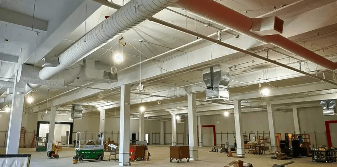 Large industrial space with exposed ceiling ducts, columns, and scattered equipment on the floor.