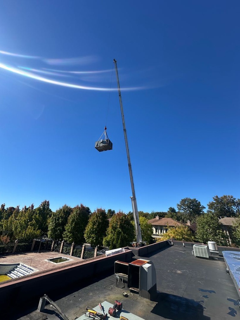A crane on a rooftop lifting an HVAC unit while five workers observe during sunset.