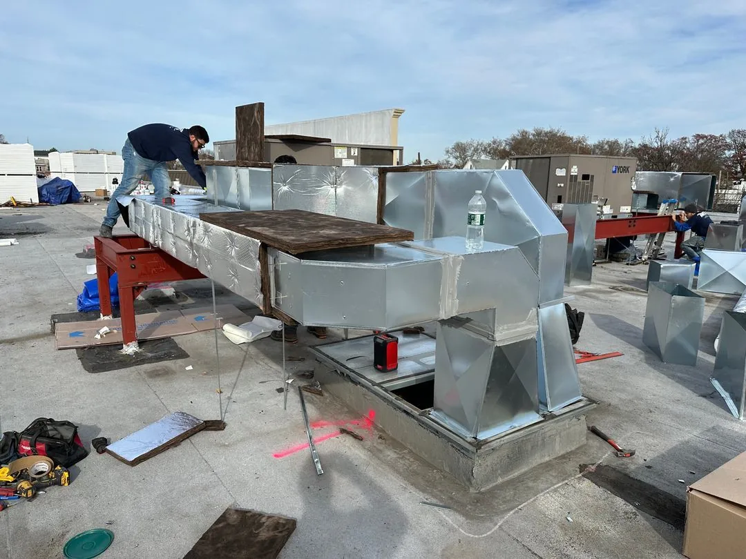 Two workers installing large silver metal air ducts on a flat rooftop under a partly cloudy sky.