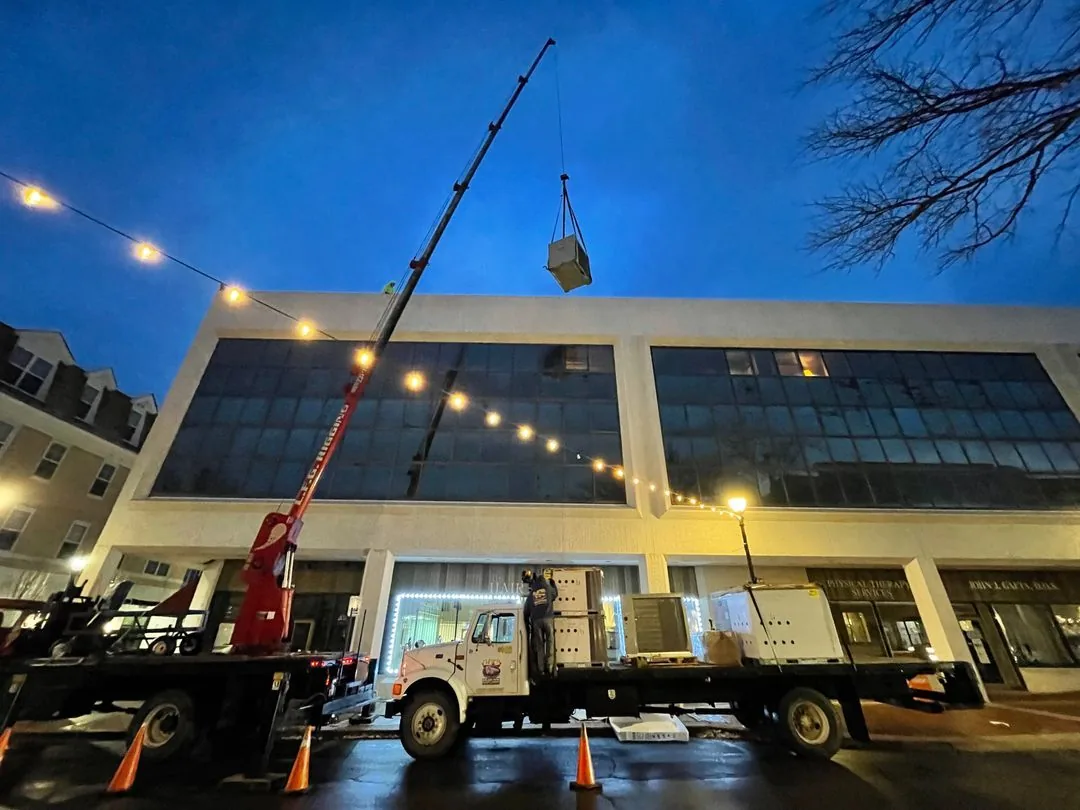 Crane lifting an HVAC unit onto a flatbed truck parked in front of a commercial building at dusk.