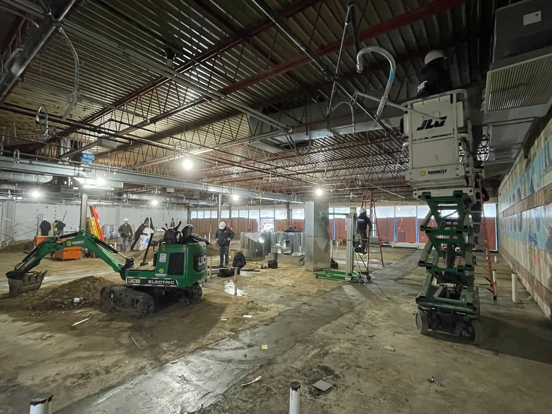 Construction workers wearing hard hats operating equipment inside a large unfinished industrial building with exposed ceiling beams and ductwork.