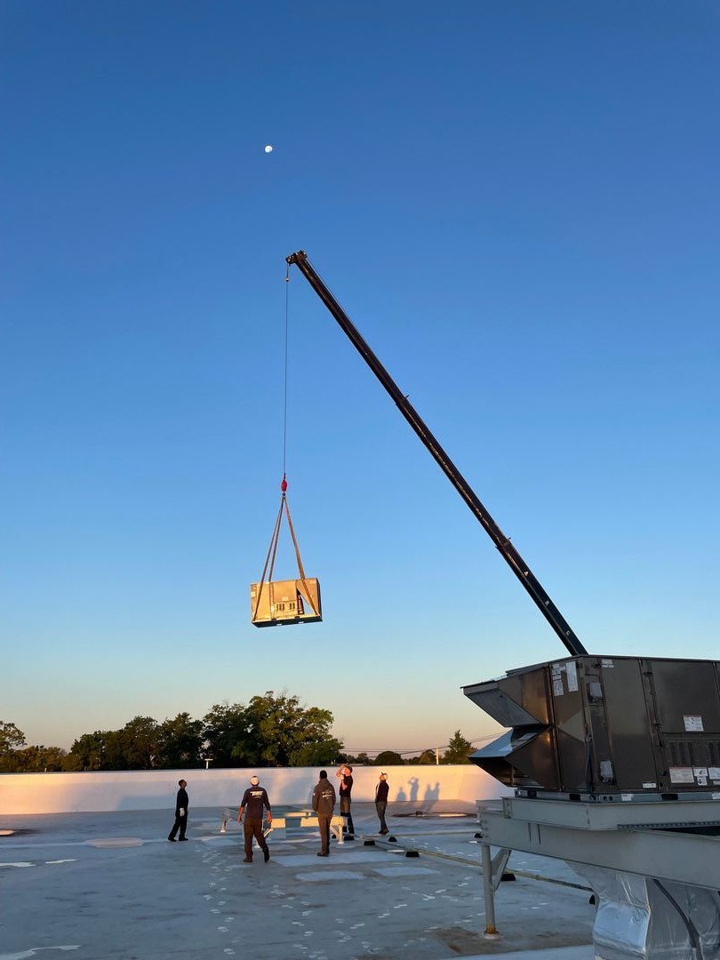 Crane lifting an air conditioning unit on a rooftop at sunset with five workers on the ground watching.