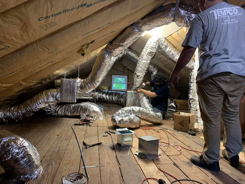 Two workers installing or repairing aluminum HVAC ductwork in an attic with exposed insulation.