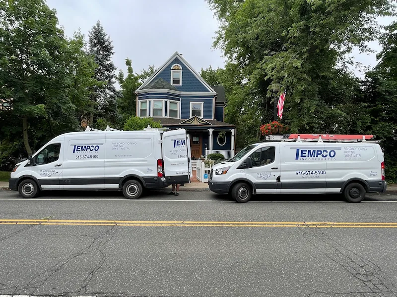 Two white TEMPCO service vans parked in front of a blue two-story house with an American flag displayed outside.