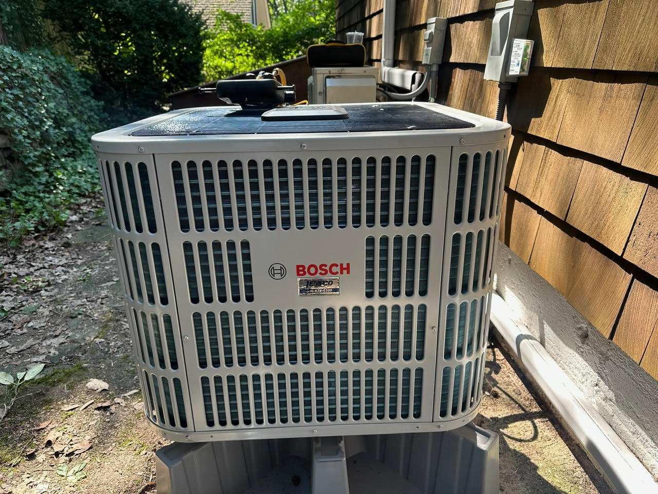 Bosch outdoor HVAC unit installed beside a wooden shingle house wall with surrounding greenery.