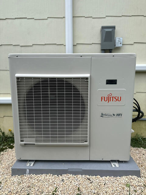 Fujitsu outdoor air conditioning unit installed on gravel beside a beige house exterior.