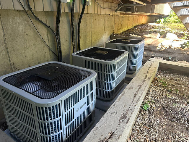 Three Bosch air conditioning units installed outdoors alongside a concrete wall and wooden border with surrounding gravel and sunlight.