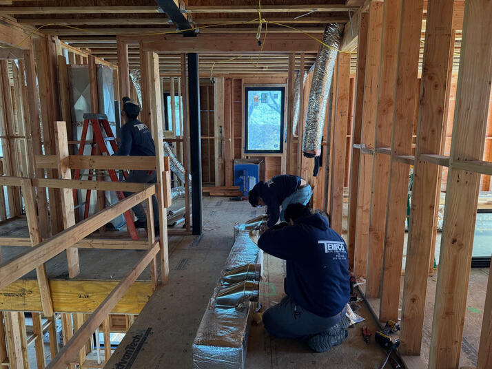 Three workers in hoodies installing ductwork inside a wooden framed house under construction.