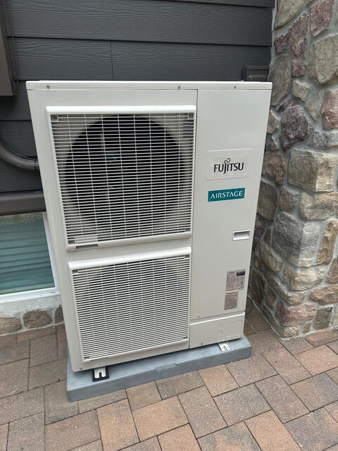 Outdoor Fujitsu Airstage air conditioning unit installed beside a stone wall and house siding.