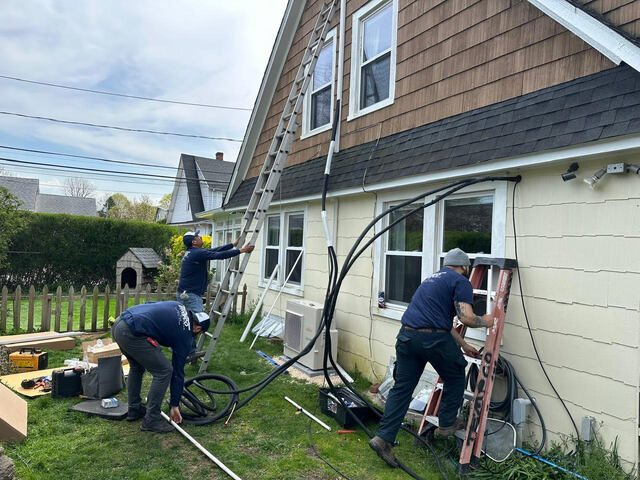 Three workers installing or repairing outdoor cables on the side of a house with ladders and tools on the ground.
