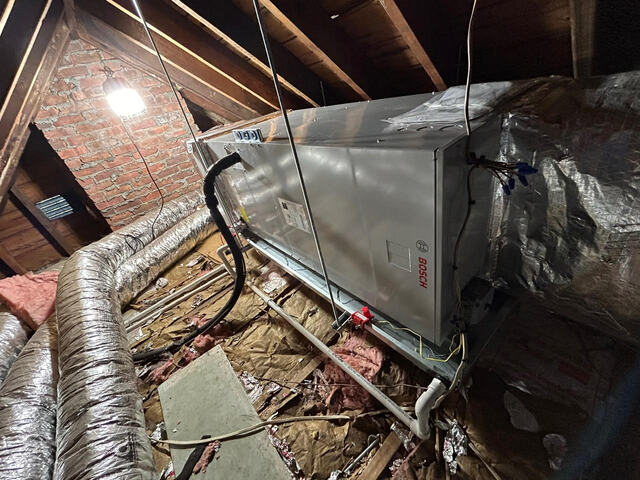 Bosch HVAC unit installed in an attic with exposed wooden beams, ductwork, insulation, and a bright light bulb.