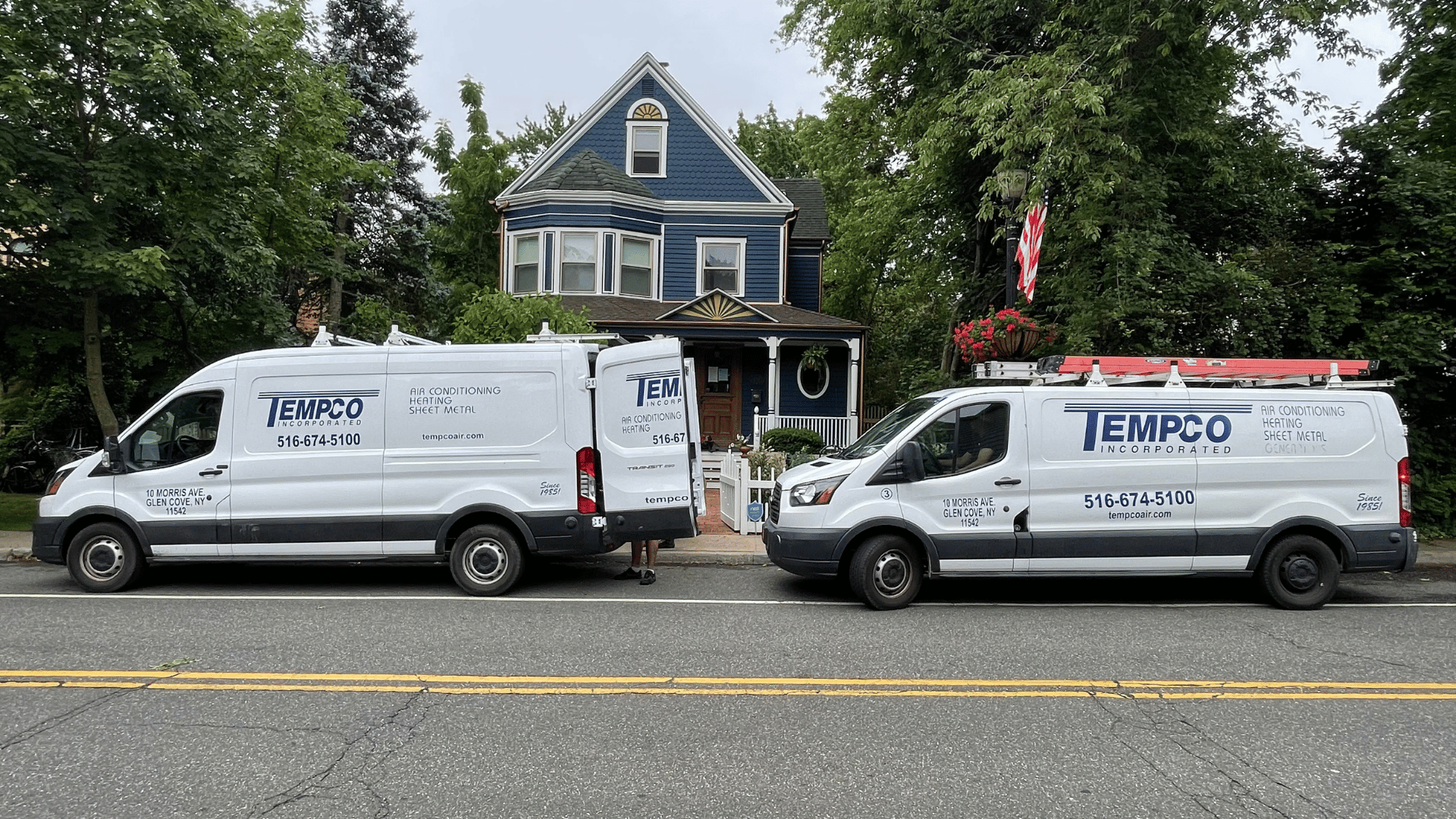 Two white TEMPCO service vans parked in front of a blue two-story house with an American flag displayed outside.