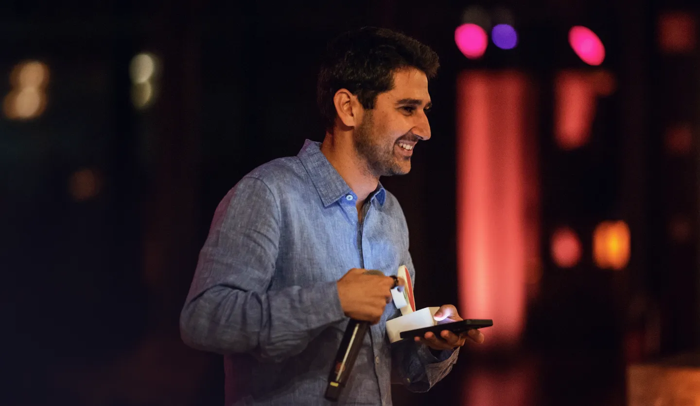 Kian Katanforoosh smiling on stage, holding a microphone and phone, with colorful stage lighting in the background.
