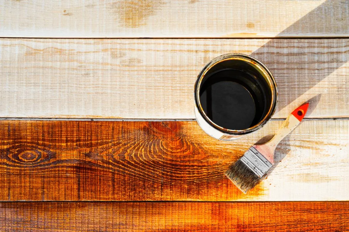 Dažų skardinė ir teptukas padėtas ant medinio, pusiau nudažyto paviršiaus.