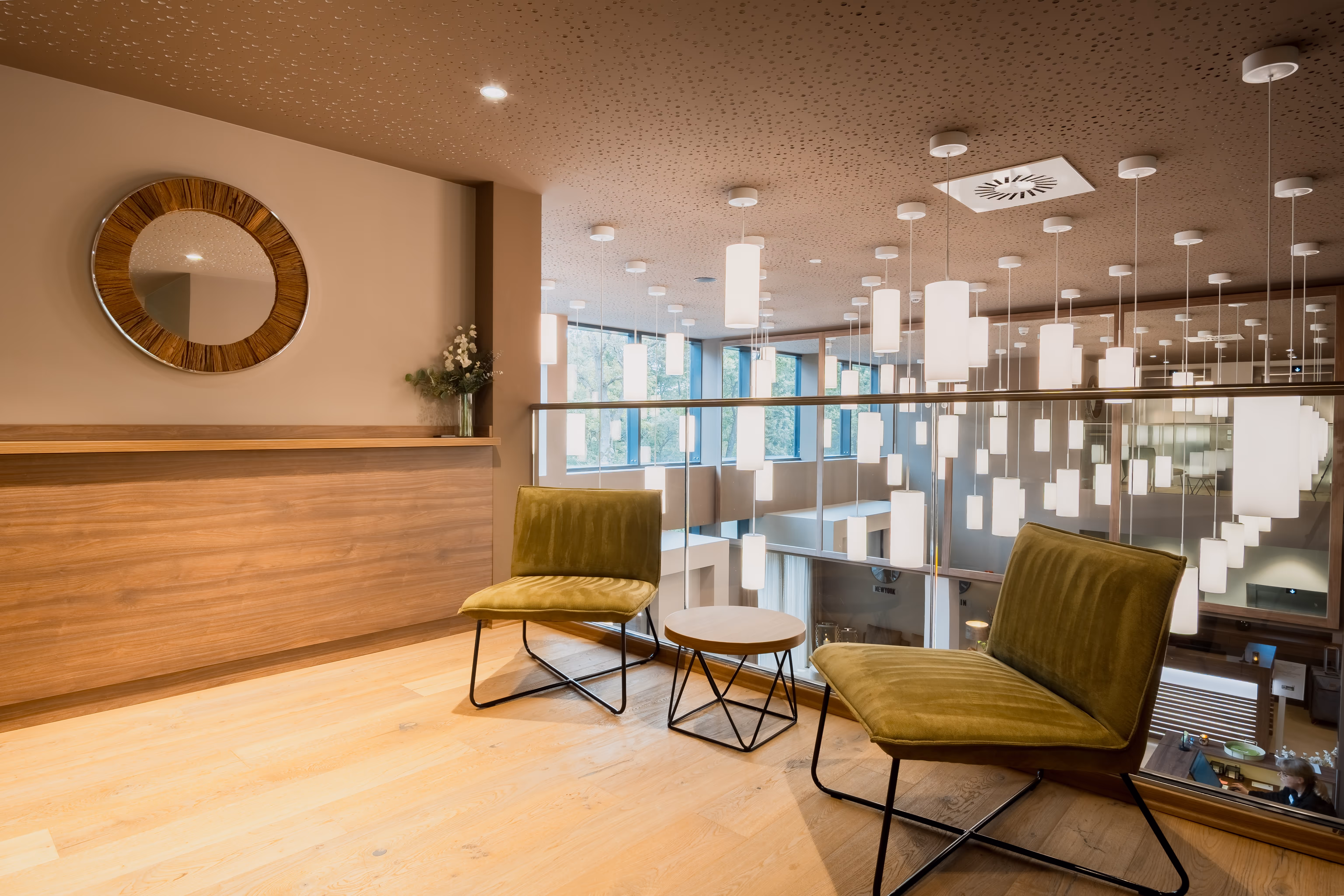 Moderner Sitzbereich mit zwei grünen Sesseln und kleinem runden Tisch vor Glasgeländer mit hängenden weißen Lampen.