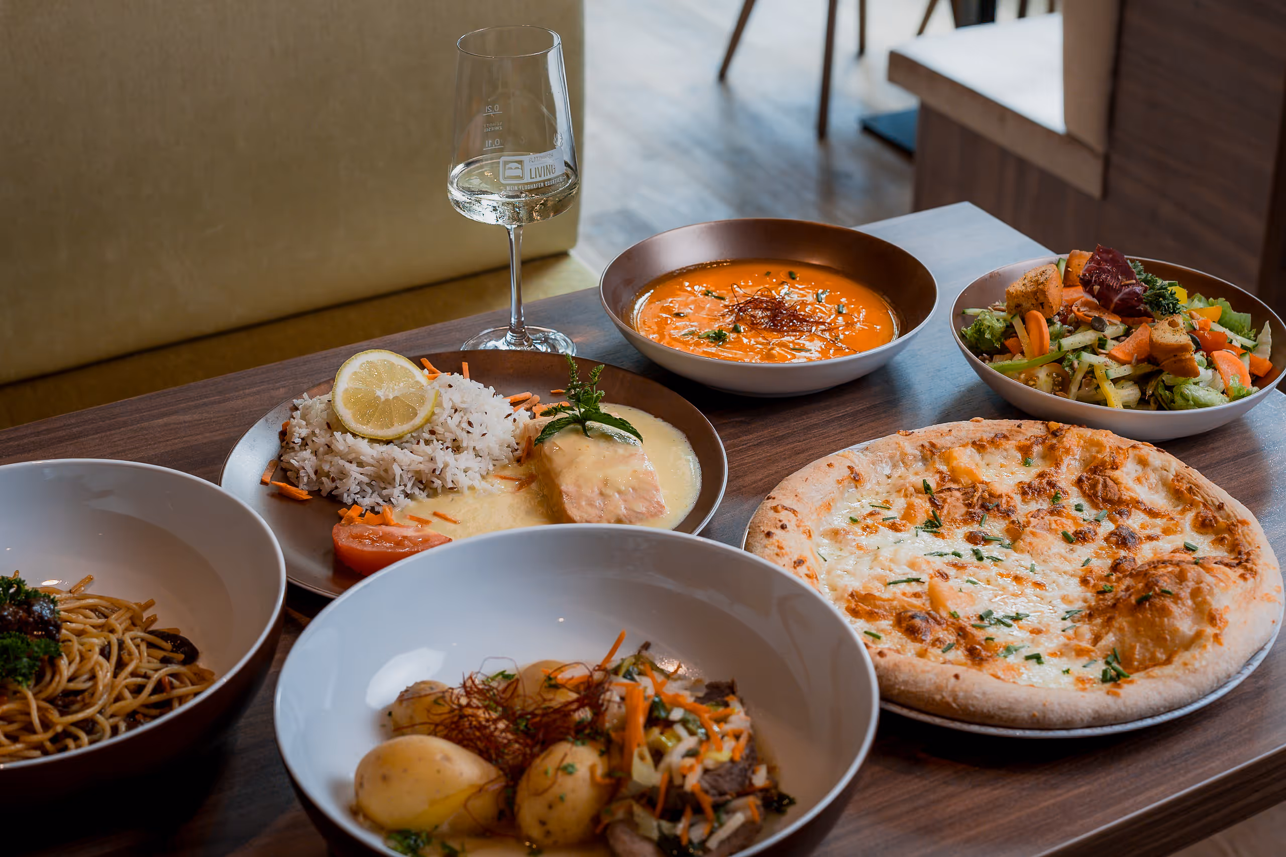 Tisch mit verschiedenen Gerichten inklusive Pizza, Salat, Suppe, Reis mit Fisch, Nudeln und einem Glas Weißwein.
