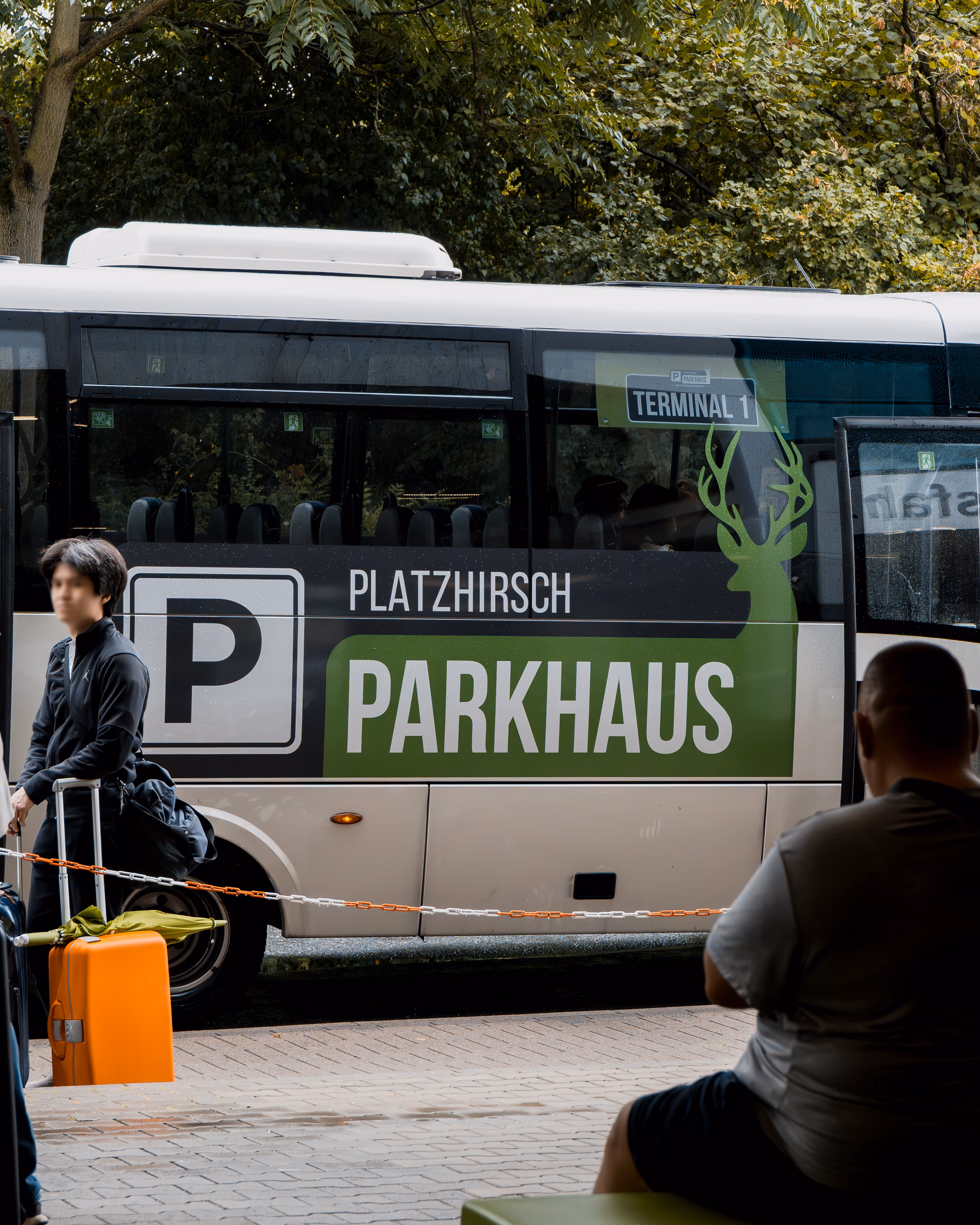Ein Parkshuttlebus mit der Aufschrift 'Platzhirsch Parkhaus' und 'Terminal 1', daneben steht ein Mann mit einem orangefarbenen Koffer.