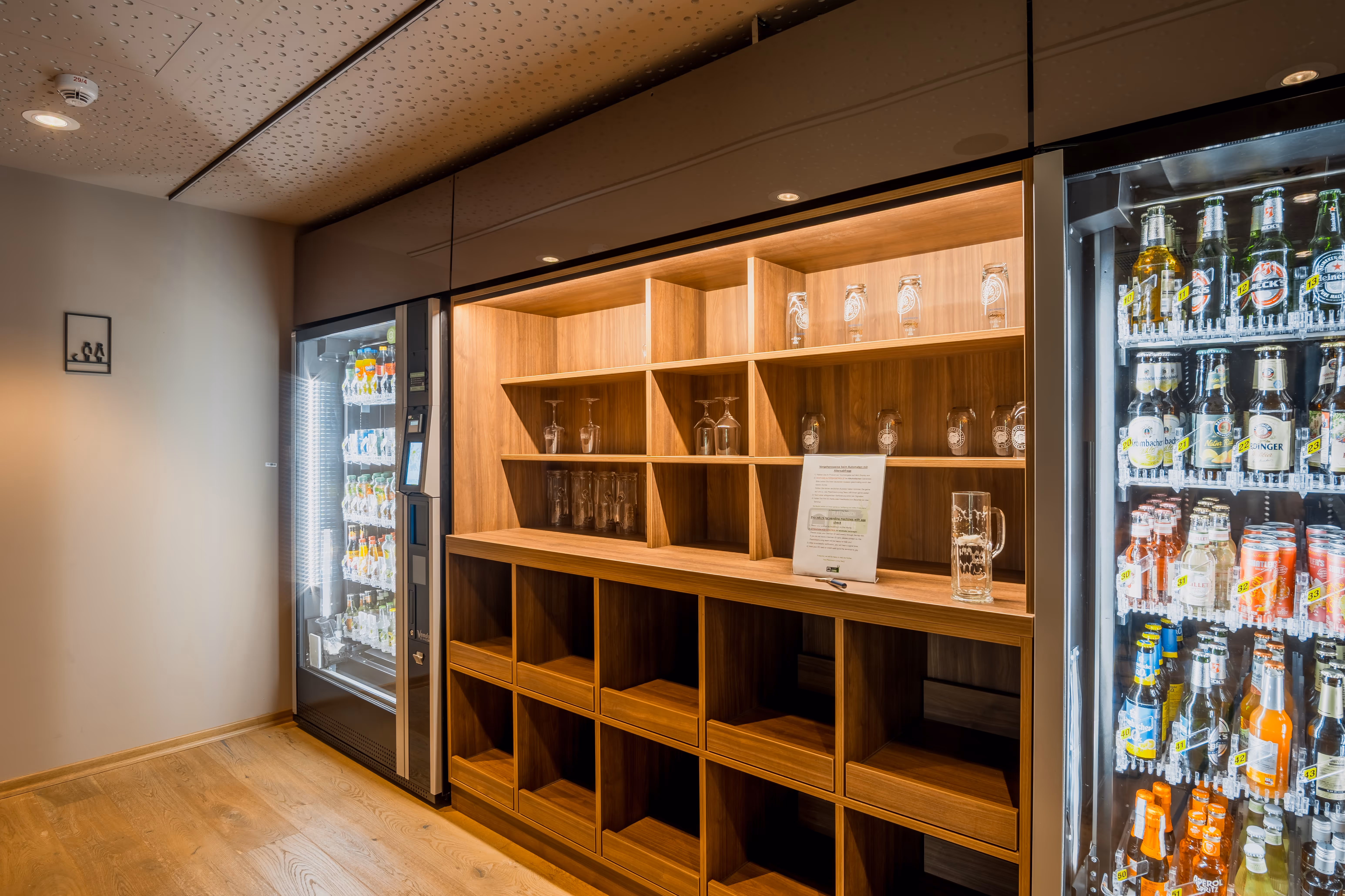 Moderner Getränkekühlschrank mit verschiedenen Flaschen und ein Holzregal mit Gläsern und einem Informationsblatt.